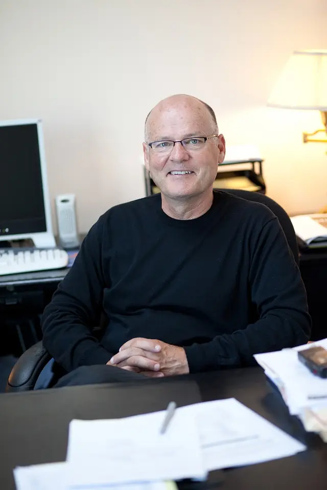 A partner in glasses and a black sweater seated at a desk with a monitor, keyboard and lamp behind him